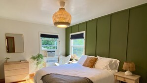 Et soveværelse med en stor seng, et natbord med en lampe, en kommode og et vindue med udsigt til grønne omgivelser.