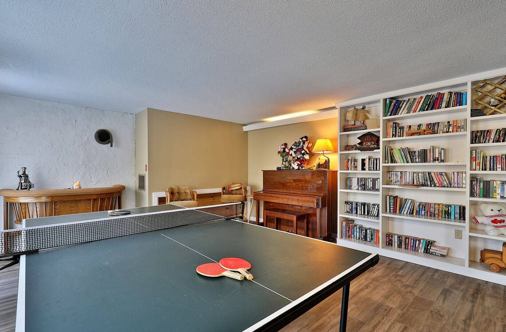 A room with a ping pong table, a piano, a bookshelf, and a sofa.