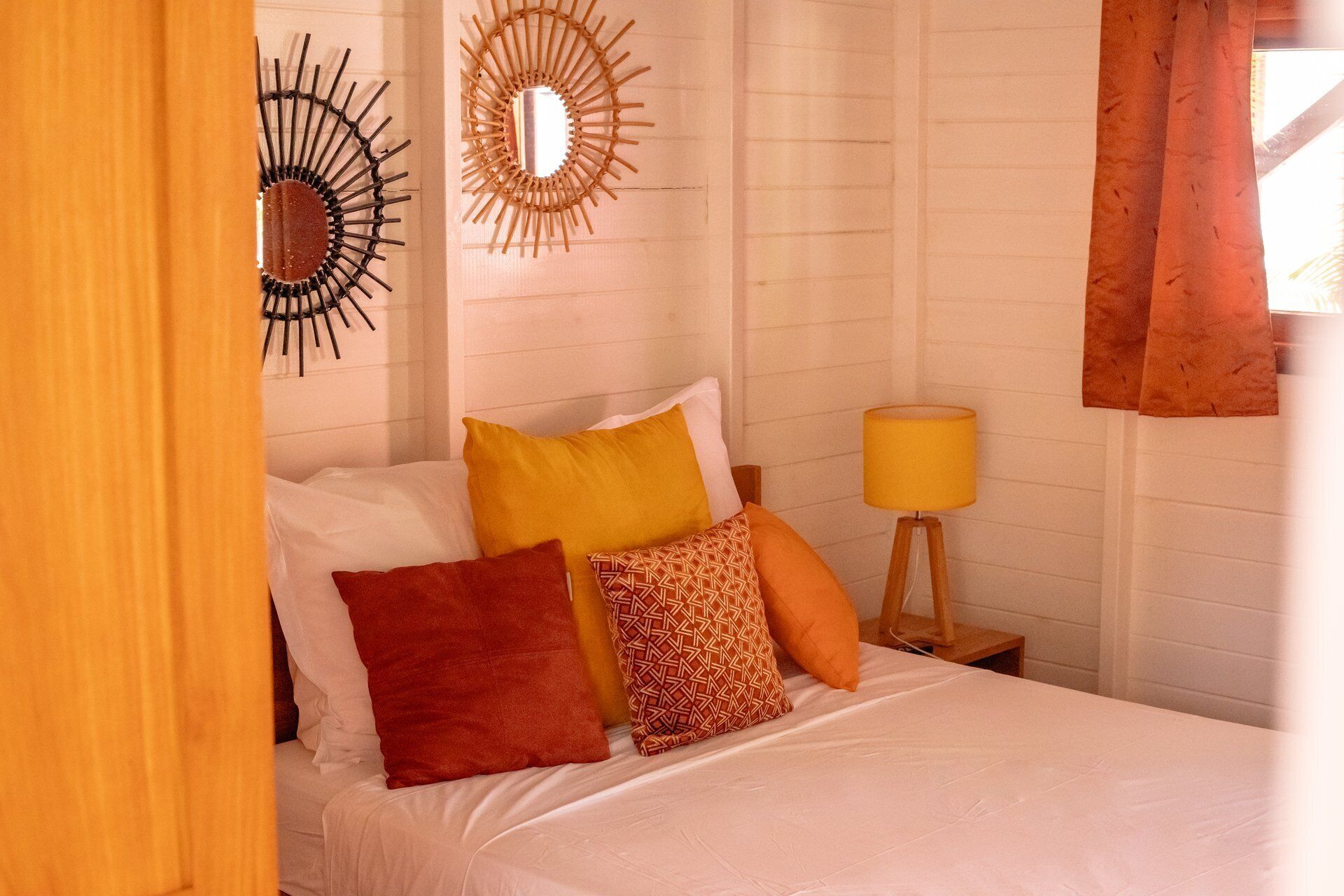 A bed with white bedding, a yellow pillow, and a patterned pillow. There is a mirror on the wall and a lamp on a bedside table.