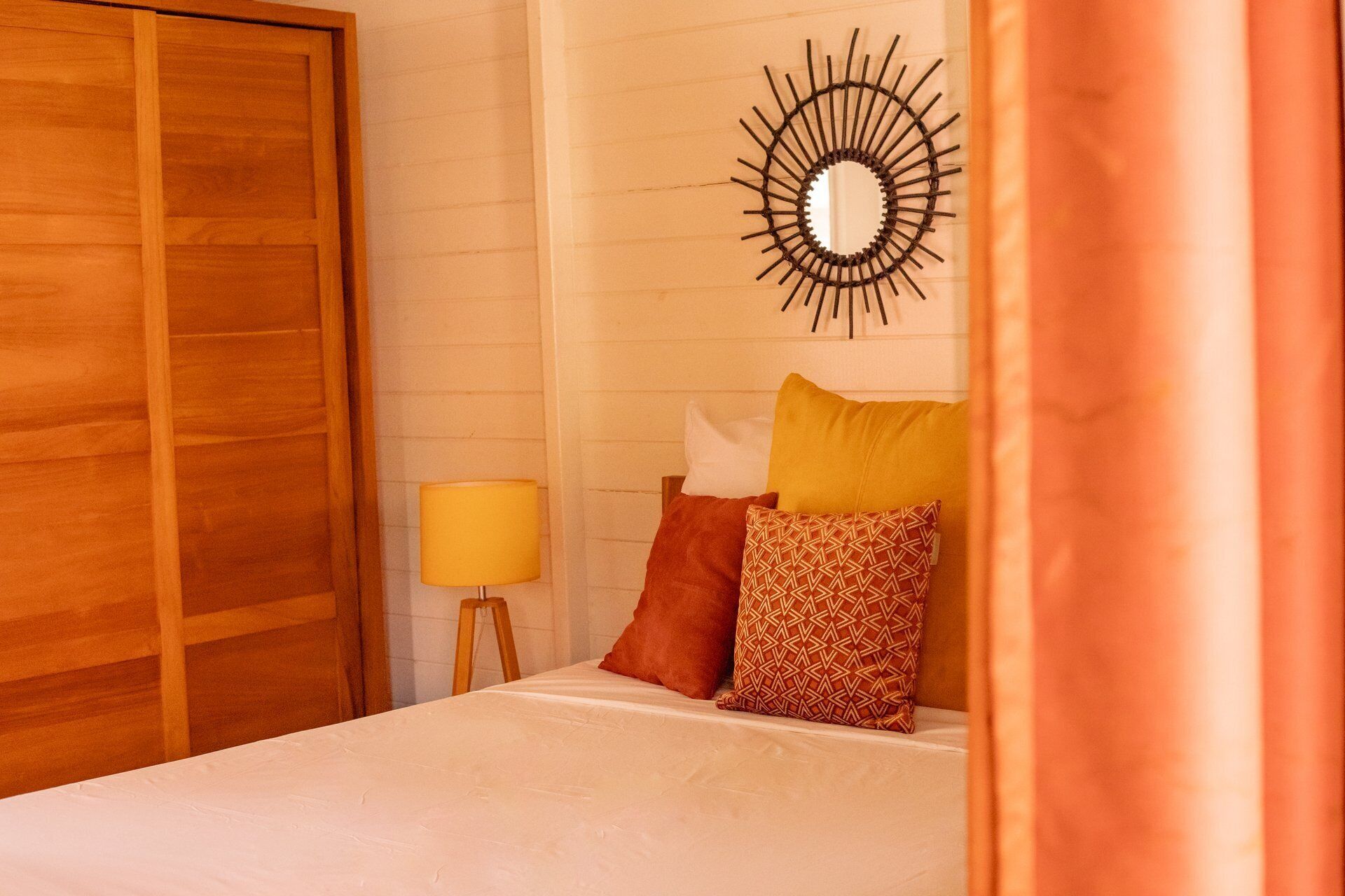 Een slaapkamer met een houten garderobe, een bed met opvallende kussens, een spiegel aan de muur en een lamp.