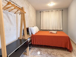 Quarto com cama, guarda-roupa de madeira, mesa de cabeceira, janela com cortinas e luminária na parede.