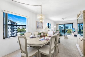 A dining area with a table set for four, a chandelier, and a view of buildings through large windows. - Estero Cove 341 (Fort Myers Beach)
