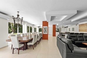 Un espace repas moderne avec une longue table en bois et des chaises blanches, un canapé en cuir noir et une grande fenêtre donnant sur un paysage verdoyant.