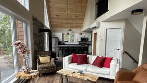 Un salón amplio con un sofá blanco, cojines rojos, una mesa de centro y un sillón con estampado. Se aprecia una cocina al fondo.