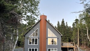 Una casa de dos pisos con una chimenea destacada, ventanas amplias y una terraza de madera.