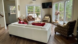Amplia sala de estar con un sofá blanco grande, dos sillones estampados, techo de madera y ventanas grandes que ofrecen vistas al exterior.