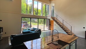 Une cuisine moderne avec un îlot central en bois, de grandes fenêtres et un escalier menant à un niveau supérieur.