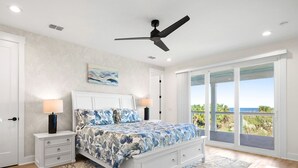A bedroom with a bed, nightstands, a ceiling fan, a large window with a view, and a door. - Beachside Beauty (Palm Coast)