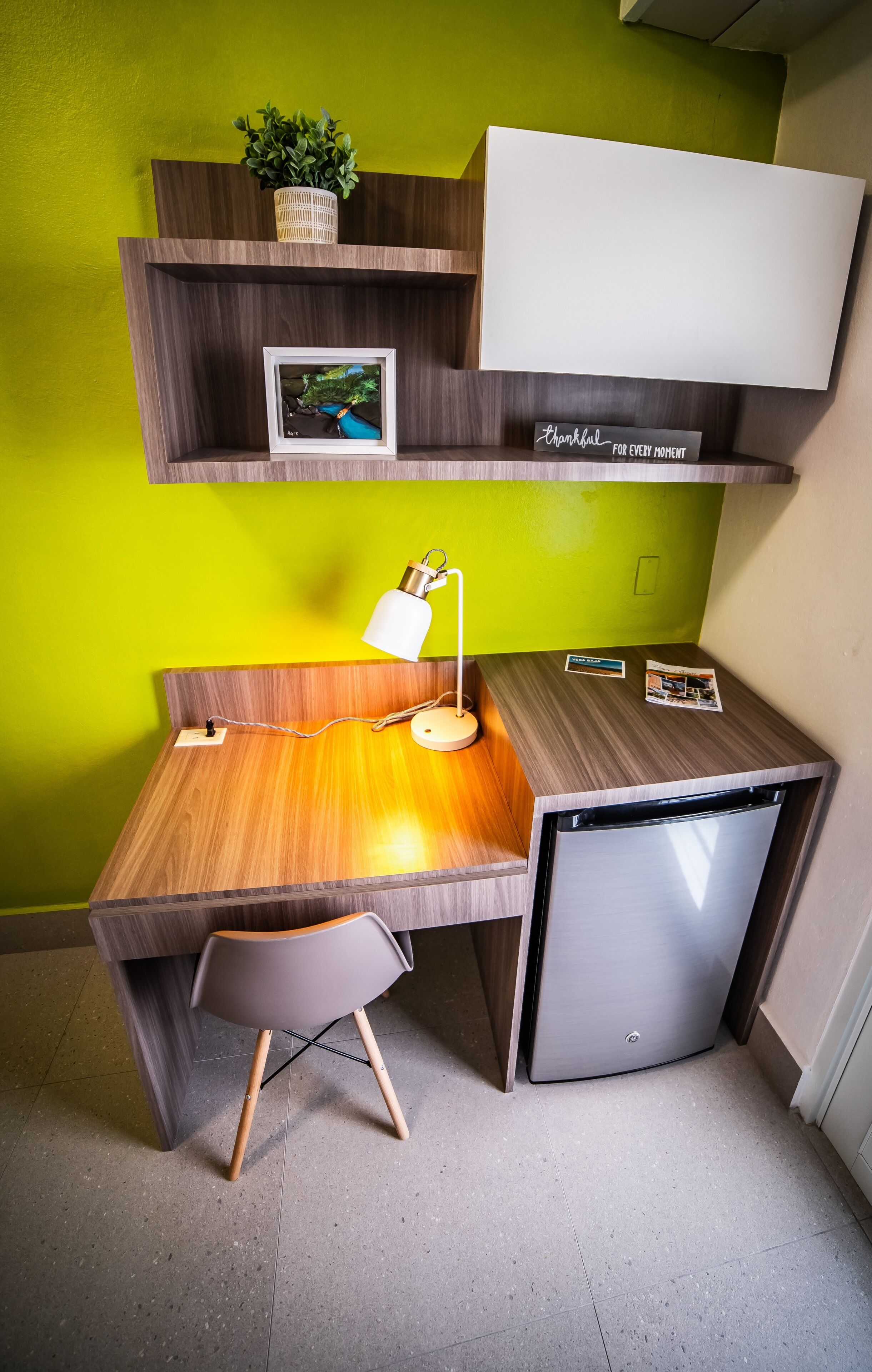 Una kitchenette compacta con encimera de madera, un refrigerador pequeño, un escritorio con silla, un estante en la pared con una planta y un cuadro enmarcado.