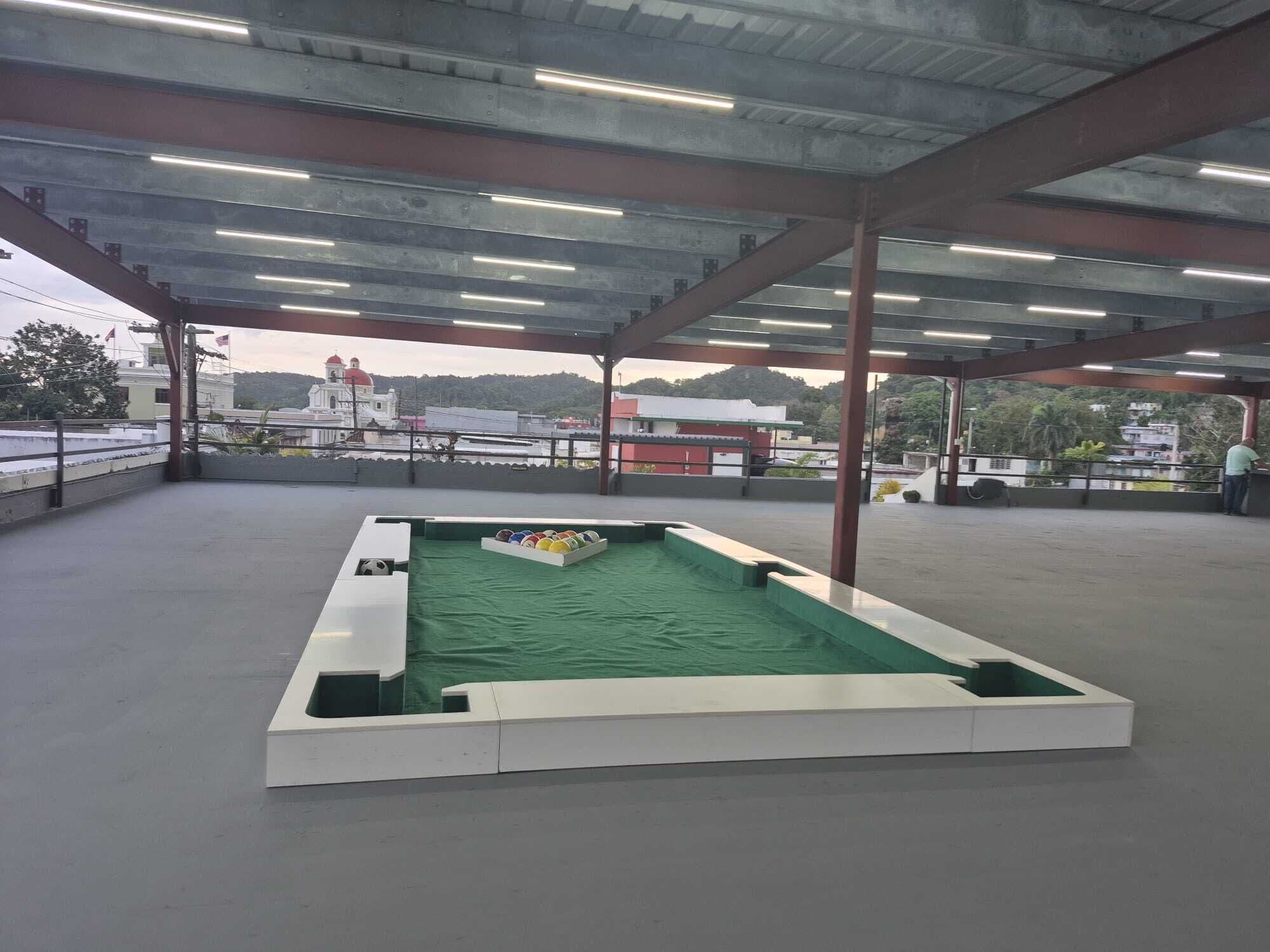 Pista deportiva interior con superficie verde, rodeada por un área cubierta con vigas metálicas e iluminación.