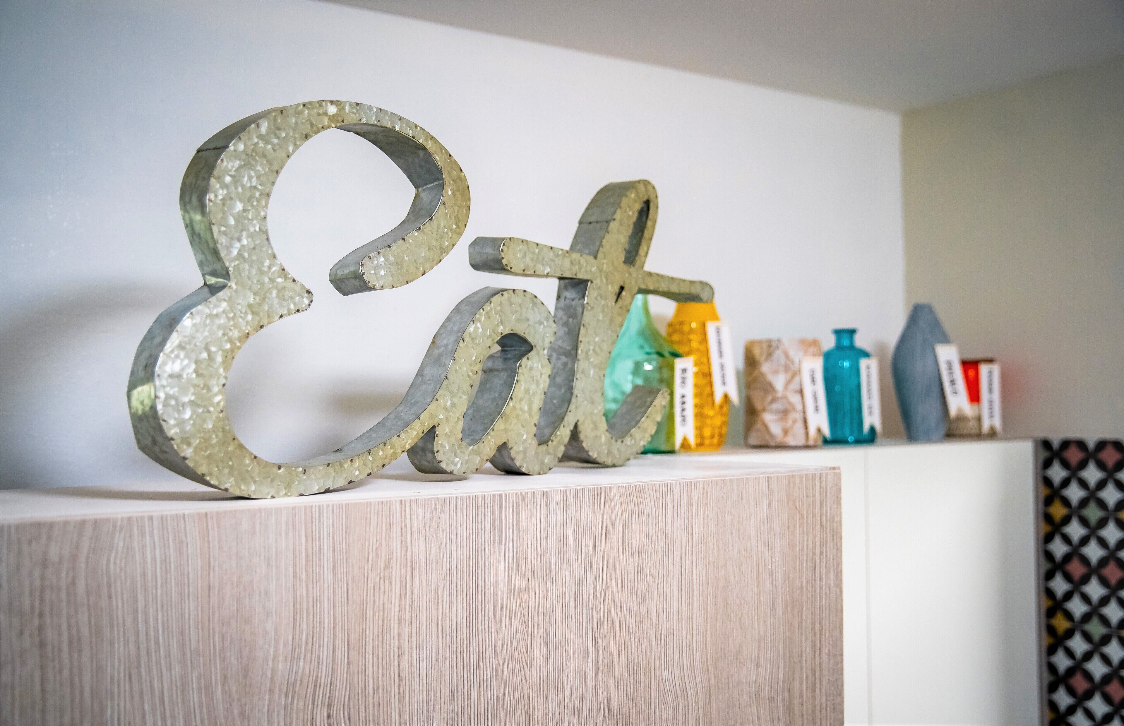 Een decoratief bord waarop 'Eet' staat, op een plank met verschillende spullen erachter.
