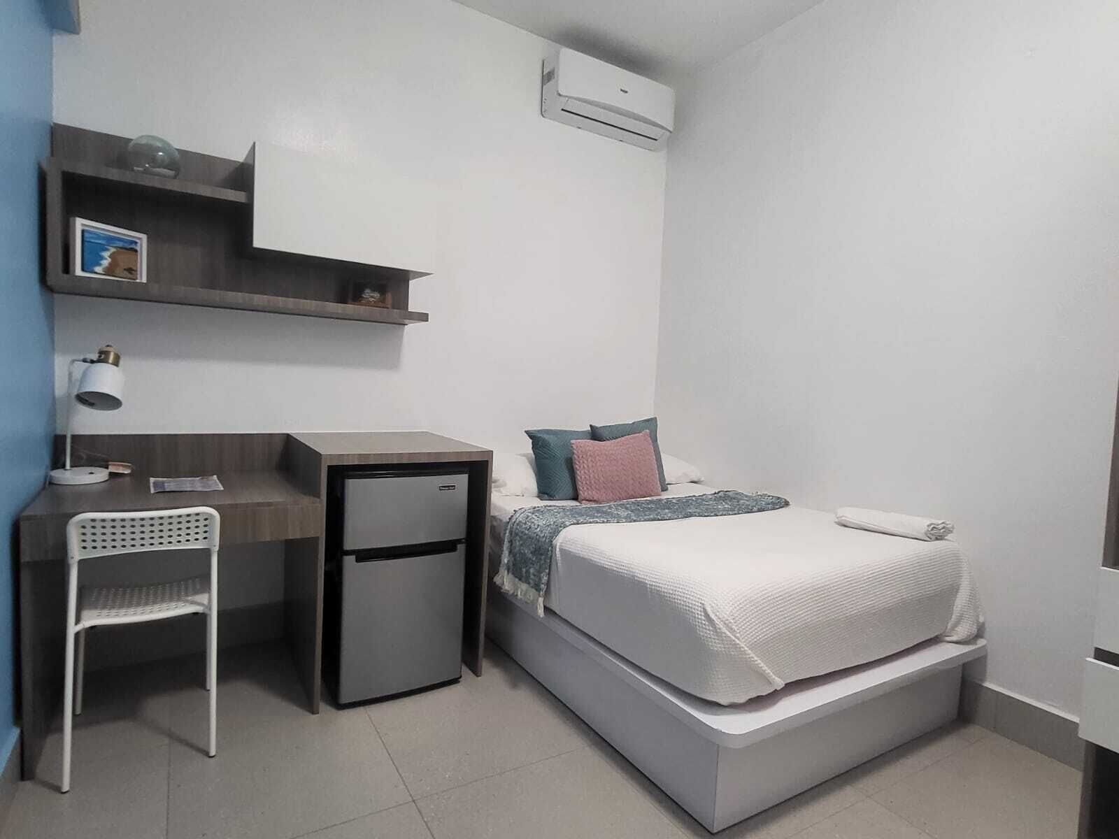 A compact hotel room with a bed, a desk, a chair, a small refrigerator, and a shelf with a picture.