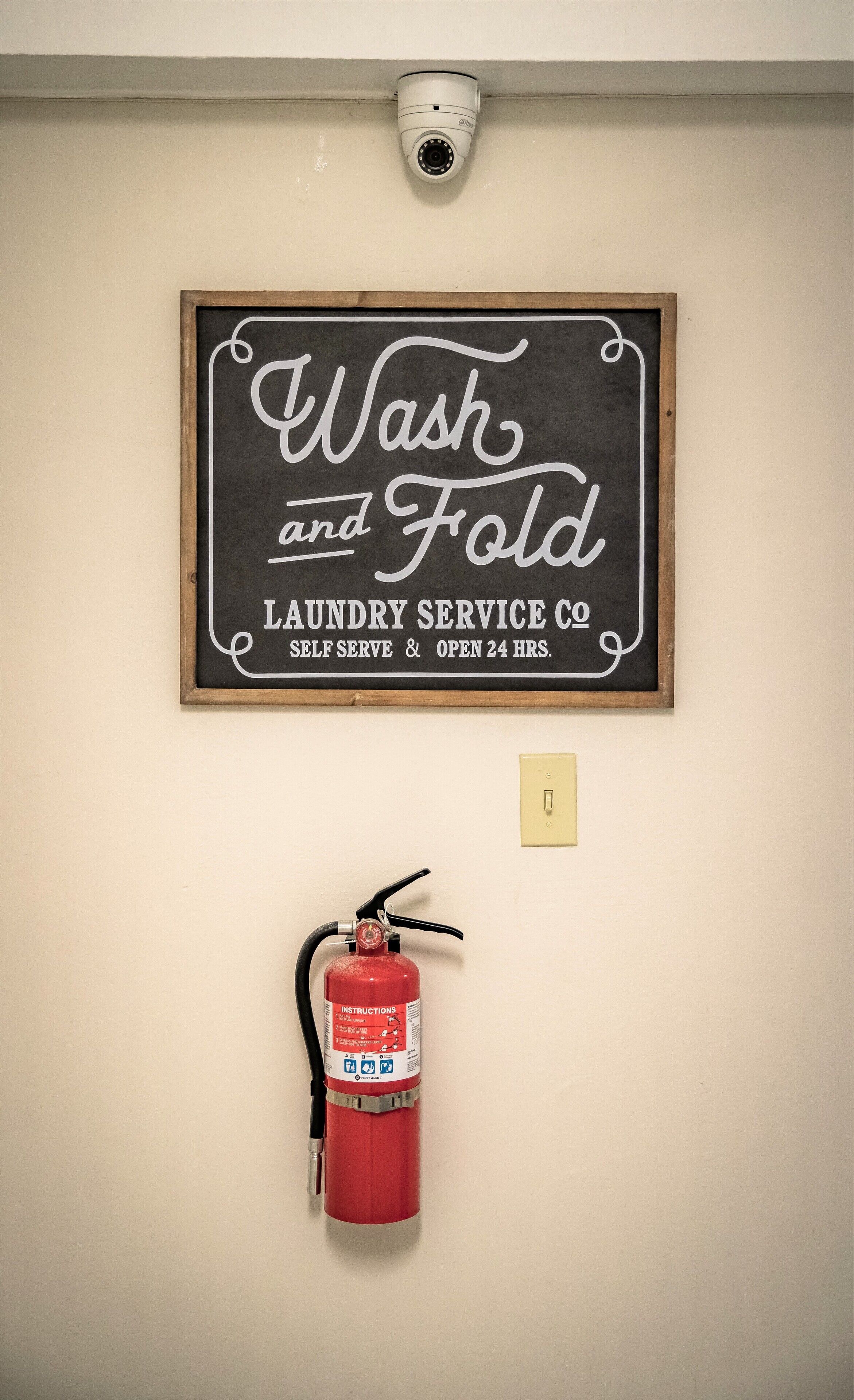 A wall-mounted sign for a self-serve laundry service, a security camera, and a fire extinguisher.