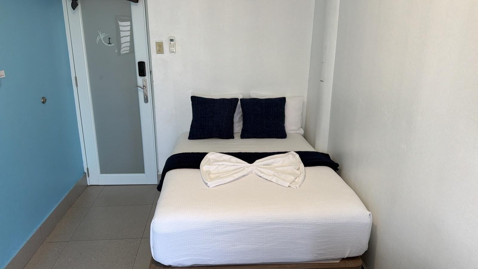 A small, single-bed room with a white mattress, two dark pillows, and a white bow on the bed. A door with a handle and a light switch is visible on the left.