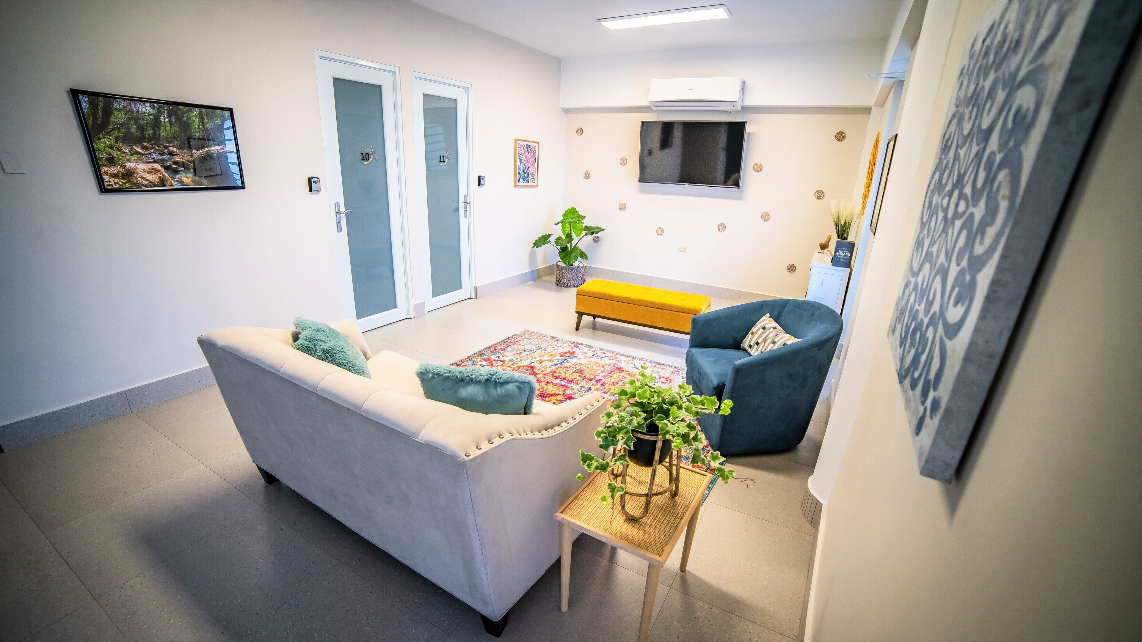 A living room with a sofa, armchair, and a small table with a plant. There are two doors, a bench, and a TV mounted on the wall.