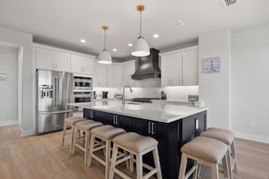A modern kitchen with stainless steel appliances, a central island with stools, pendant lighting, and a large range hood.