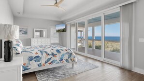A bedroom with a large bed, a ceiling fan, a TV, a dresser, and a view of the ocean through large sliding doors. - Serenity Shores (Palm Coast)