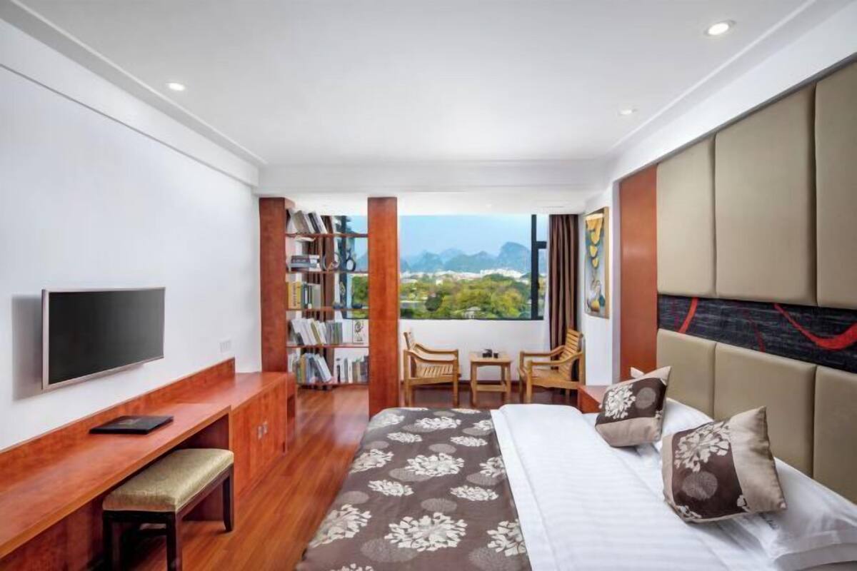 Ein Hotelzimmer mit Bett, Schreibtisch, Fernseher, Bücherregal und Blick ins Freie.