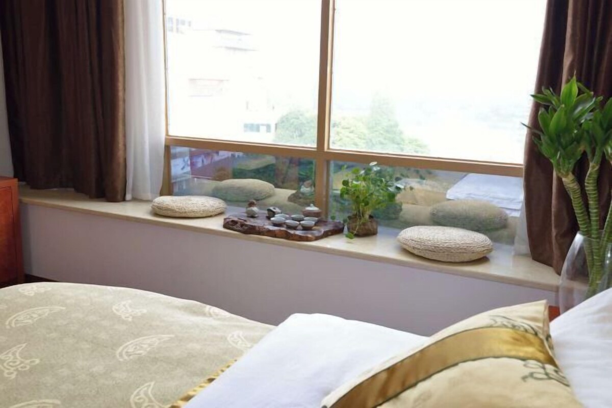 A room with a bed, a window with a view of the outside, and a small shelf with decorative items.