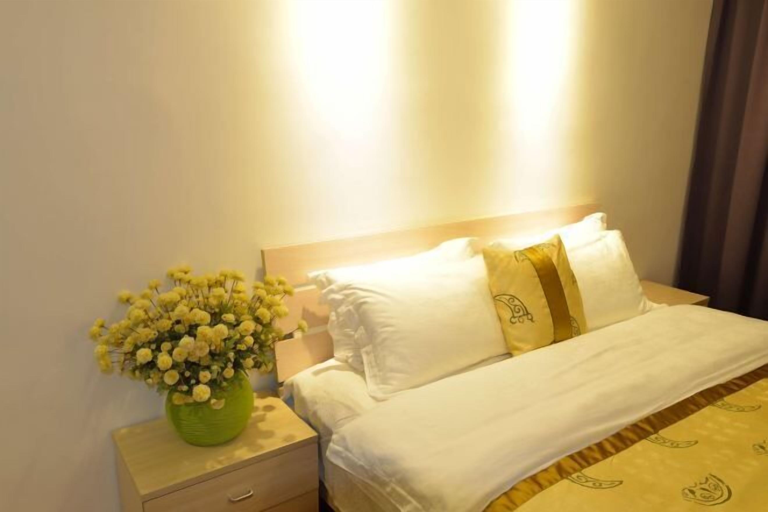 A neatly made bed with white bedding and a yellow patterned throw. A green vase with yellow flowers is placed on a bedside table.