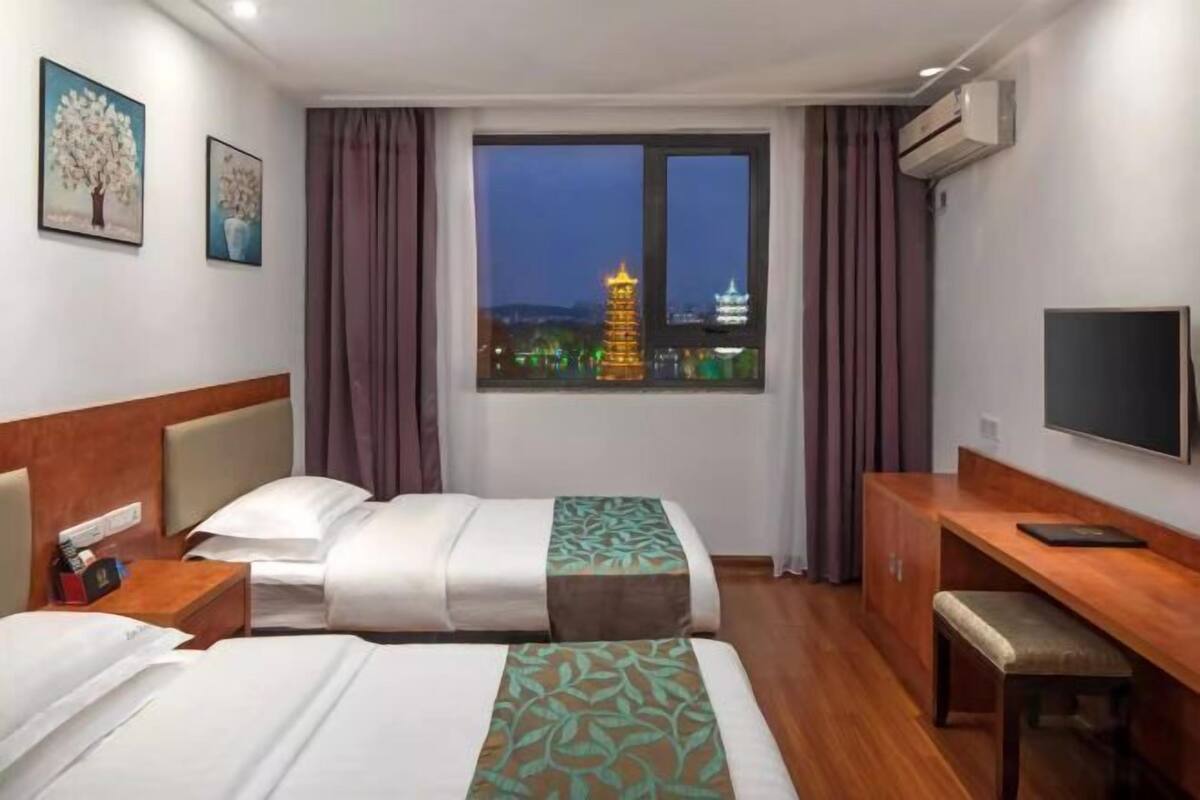 Ein Hotelzimmer mit zwei Betten, einem Schreibtisch, einem Fernseher und einem Fenster mit Blick auf die Stadt.