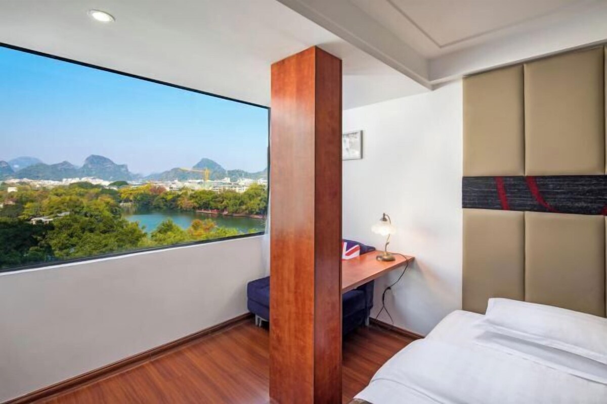 A hotel room with a large window offering a view of a lake and mountains, a wooden desk with a lamp, and a bed with a headboard.