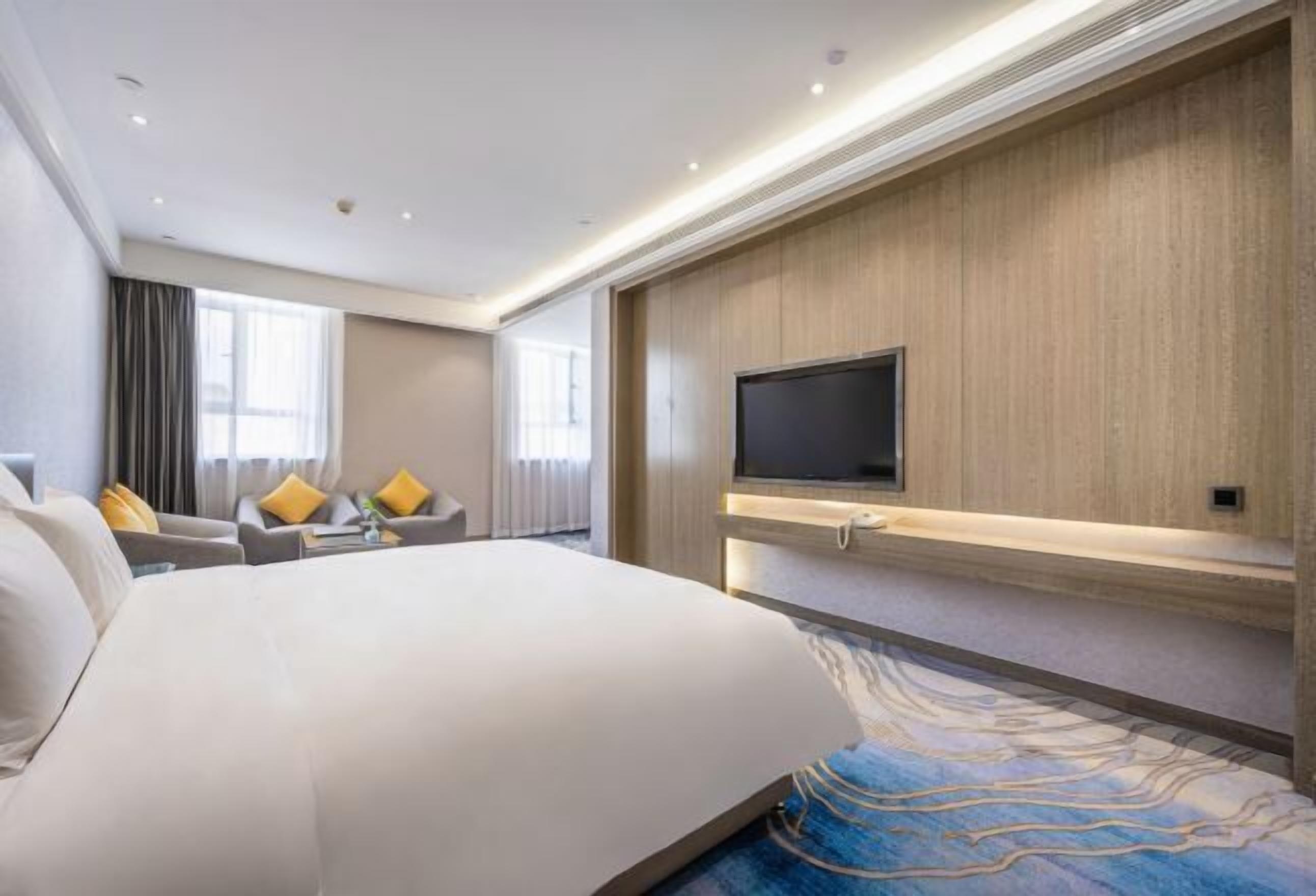 A modern hotel room with a large bed, a flat-screen TV mounted on a wooden panel, a sofa with yellow cushions, and a patterned carpet.
