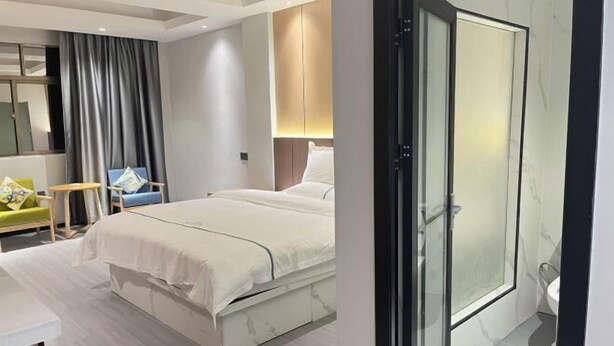 A modern hotel room with a bed, a small desk, a chair, a bathroom with a glass shower, and a large mirror. - Dongfang Guanda Business Hotel (Dongfang)