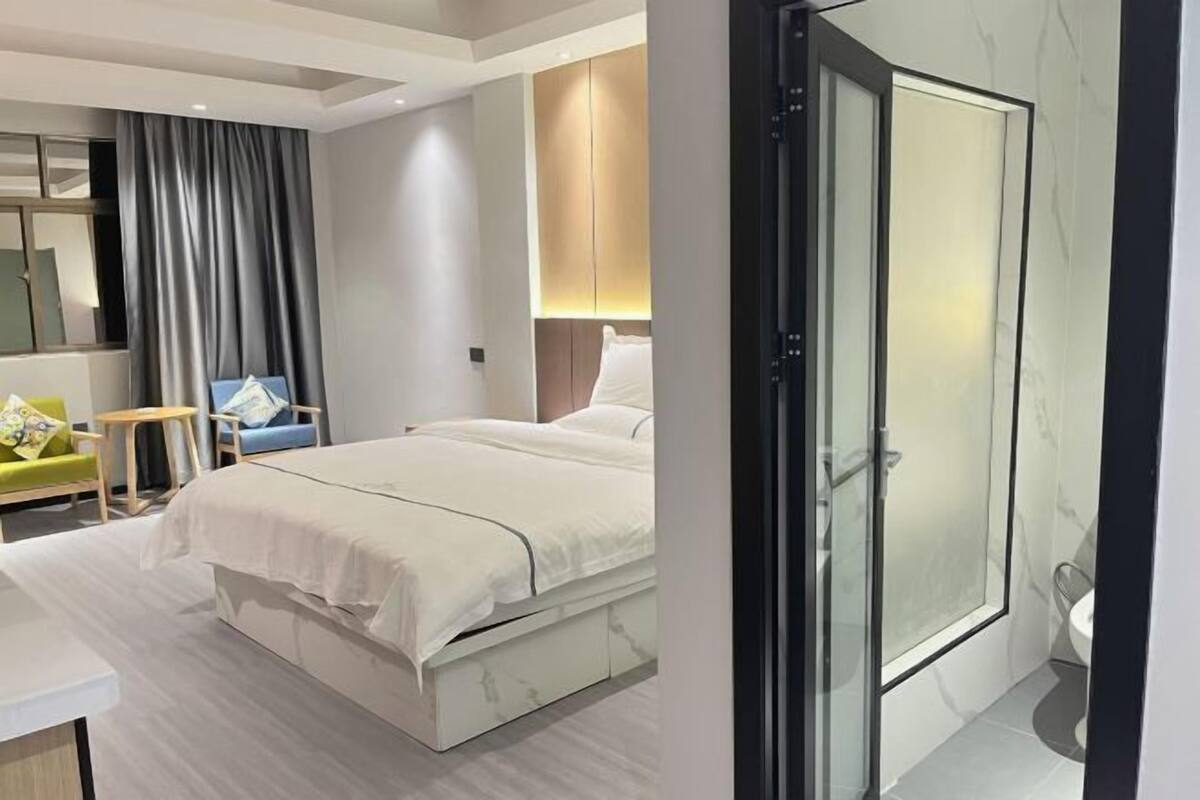 A modern hotel room with a bed, a small desk, a chair, a bathroom with a glass shower, and a large mirror.