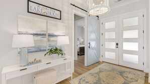 A modern entryway with a white console table, two wall-mounted lamps, a framed sign, and a large, patterned rug. - Brand New 6BR/6BA Luxury, 20min to Clemson. Wooded flat lot/firepit/pool table! (West Union)