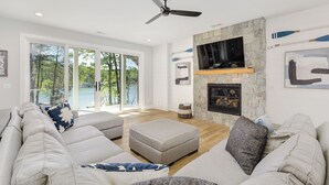 A spacious living room with a stone fireplace, a ceiling fan, and a large window offering a view of the outdoors. - Brand New 6BR/6BA Luxury, 20min to Clemson. Wooded flat lot/firepit/pool table! (West Union)