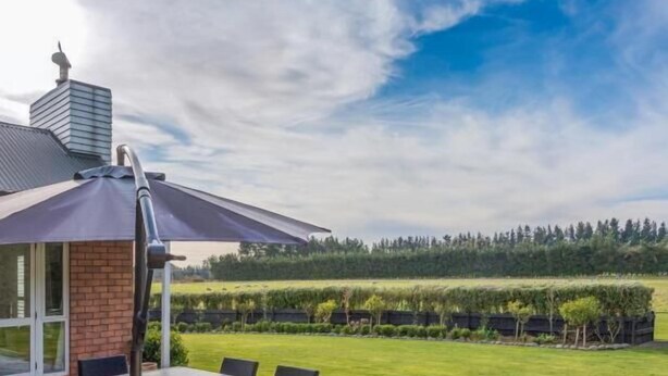 Un patio al aire libre con mesa y sillas, un paraguas grande y vistas a un campo verde y árboles.