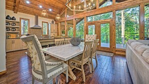 Een houten interieur met een eettafel en stoelen, een kroonluchter en grote ramen met uitzicht op het bos.