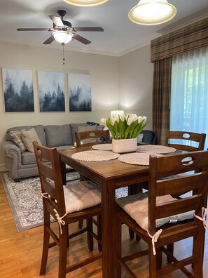 Un comedor con una mesa de madera, sillas y un ventilador de techo. Hay tres cuadros en la pared, un jarrón con flores blancas y una ventana con cortinas.