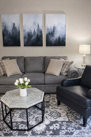 A living room with a grey sofa, a black leather armchair, a white vase with flowers, and three framed forest paintings on the wall.