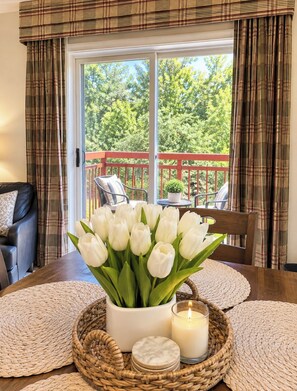 A dining area with a table set for two, a vase of white tulips, a candle, and a view of a red deck and greenery outside. - Premier RiverStone Resort&Spa•24HR Pool•Lazy River•Dollywood•By Parkway (Pigeon Forge)