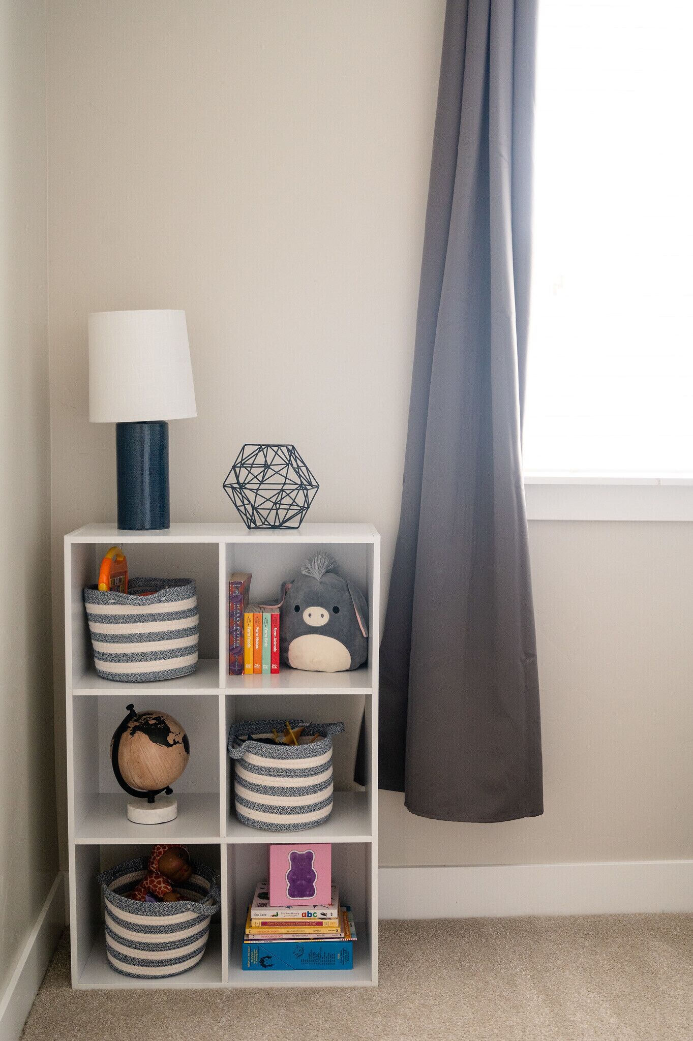 Un estante blanco con cestas de almacenamiento, un globo terráqueo, libros y un peluche, todo frente a una pared con ventana y cortinas.