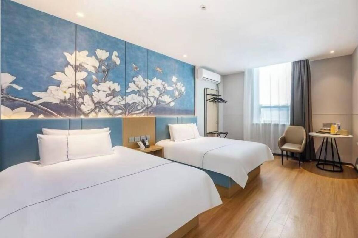A hotel room with two beds, a large wall painting, a small table, a chair, and a window with curtains.