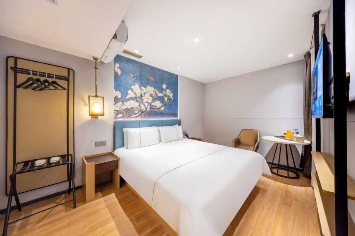 A hotel room with a bed, a desk, a chair, a coat rack, and a wall with a floral painting.