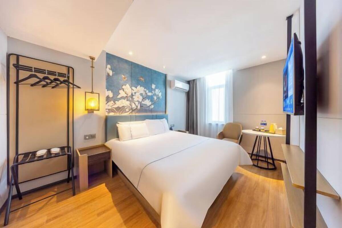 A hotel room with a bed, a desk, a chair, a TV, a coat rack, and a painting on the wall.