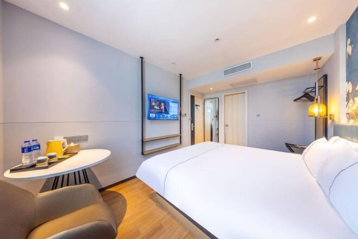 A hotel room with a bed, a desk with a chair, a TV mounted on the wall, and a door leading to another room.