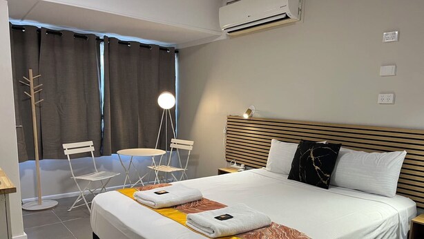 Quarto de hotel com cama, mesas de cabeceira, escrivaninha, cadeira, mesinha e janela com cortinas.