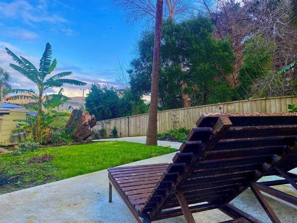 Un patio trasero con una terraza de madera, un banco y diversas plantas y árboles.