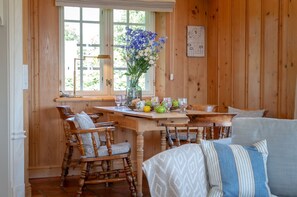 A wooden interior with a dining table set for four, surrounded by wooden chairs. A vase with flowers and fruits is on the table.