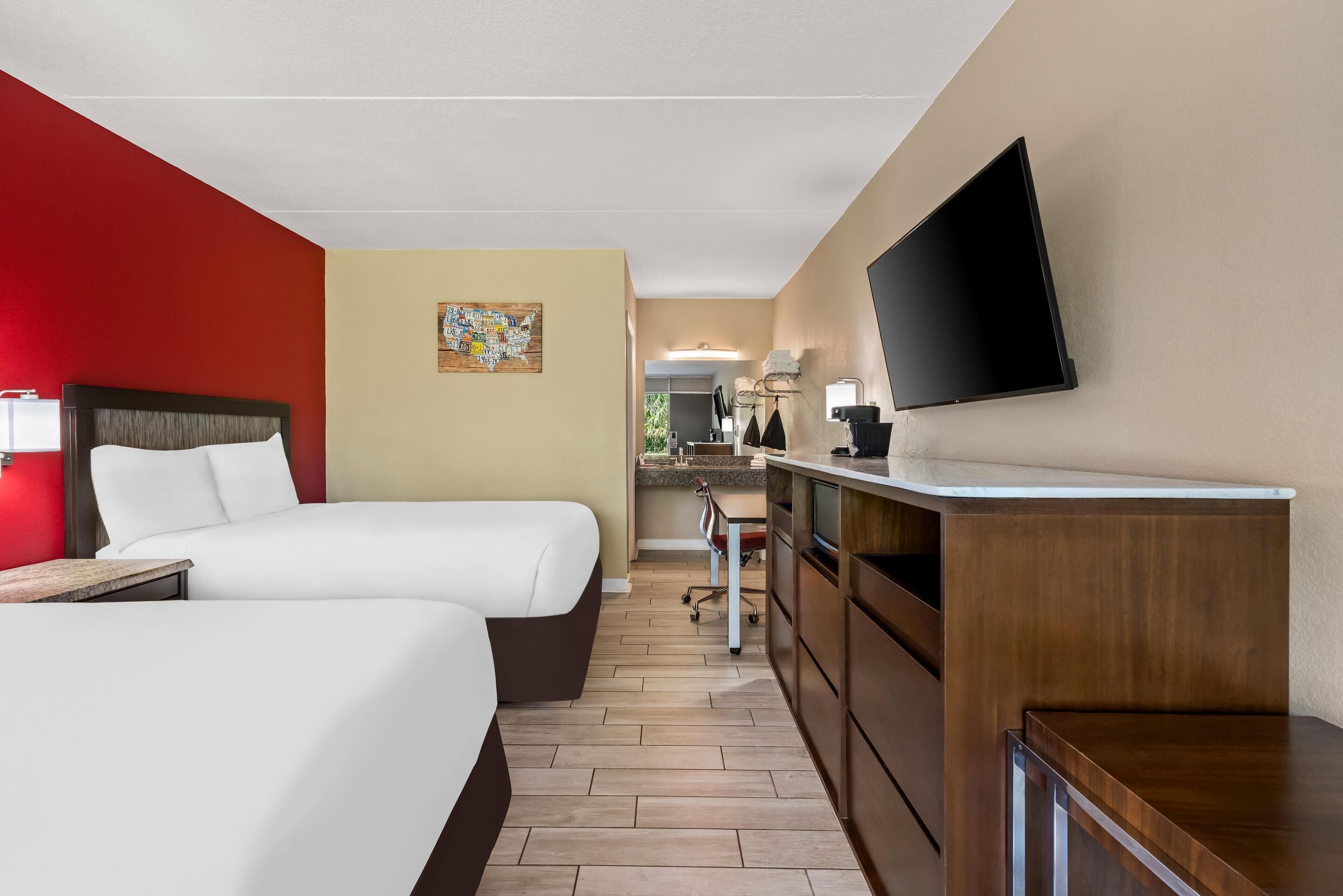A hotel room with two beds, a TV, a desk, and a kitchenette.