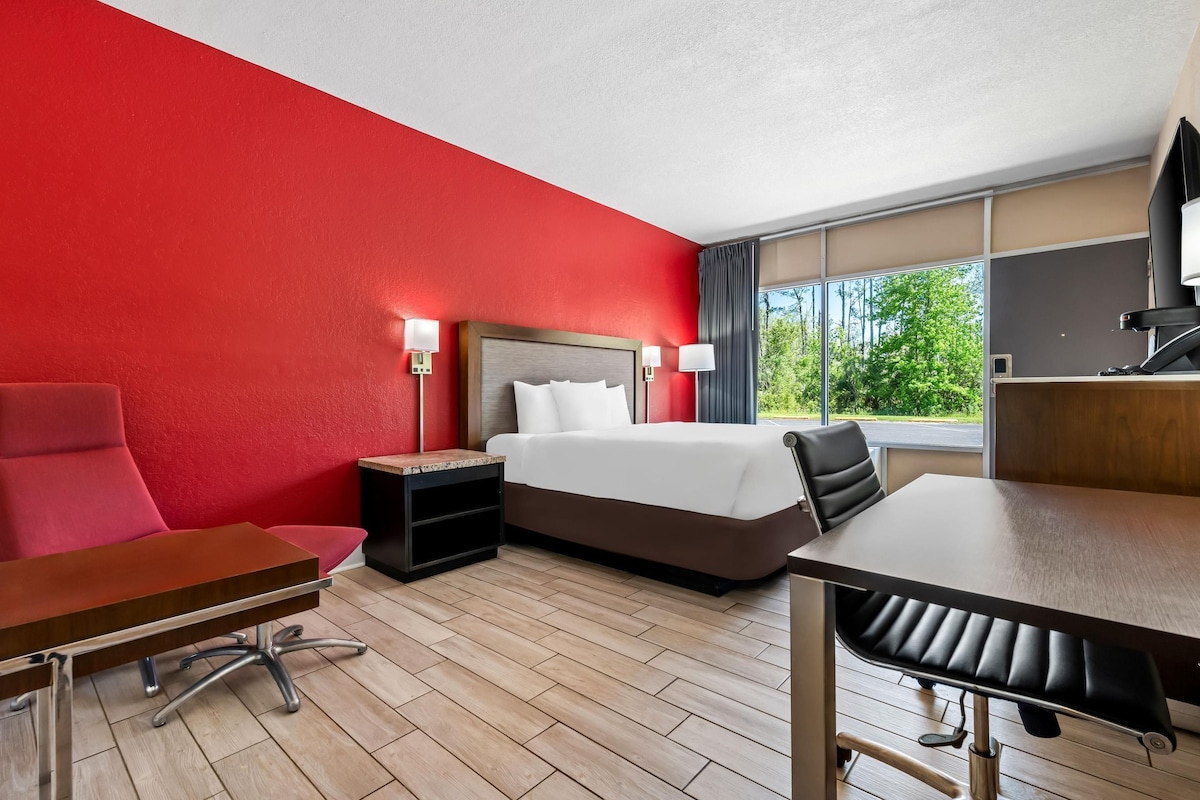 A hotel room with a bed, a desk, a chair, a nightstand, and a window with a view of trees.