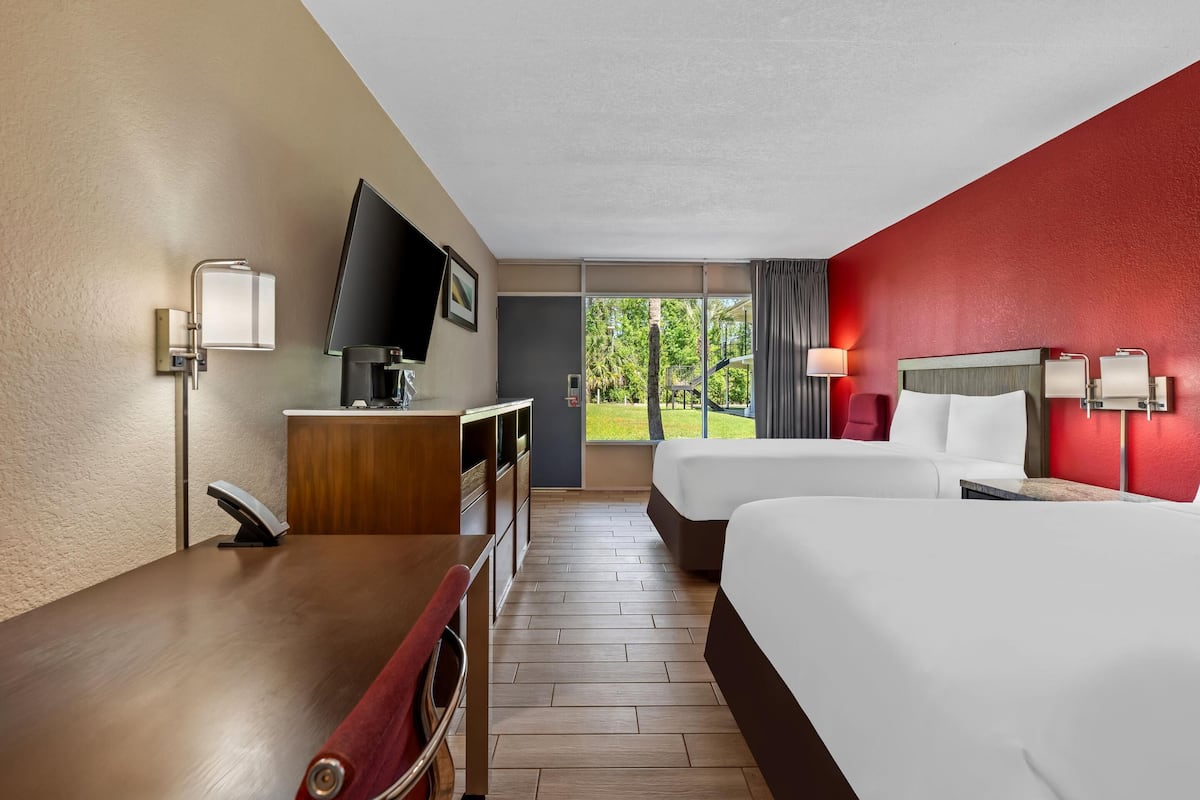 Ein Hotelzimmer mit zwei Betten, einem Schreibtisch, einem Fernseher und einem Fenster mit Blick auf Grünflächen.