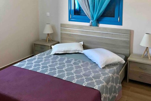 A bedroom with a bed, two bedside tables, a nightstand, a window with curtains, and a wall-mounted lamp.