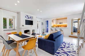 A modern living room with a sofa, dining table, and chairs. There is a kitchen area with a microwave and a TV.