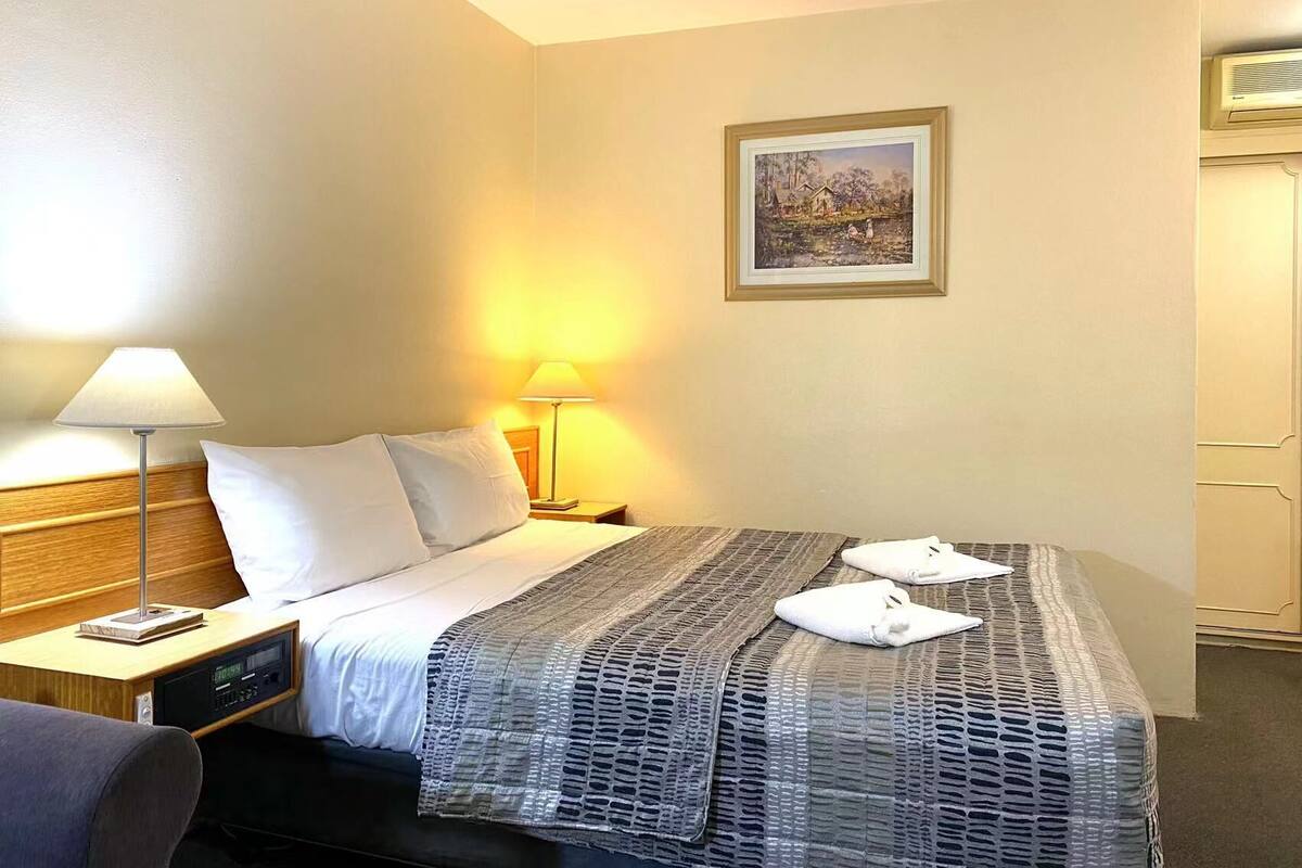 Ein Hotelzimmer mit Bett, Nachttischlampen, einem Nachttisch, einem gerahmten Bild an der Wand und einer Tür.